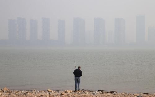 10月25日,一名男子在長沙湘江畔釣魚,江對面一片建筑在霧霾籠罩下若隱若現(xiàn)。新華社記者 李尕 攝